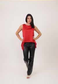 Top senza maniche in raso rosso con un orlo fluente abbinato a pantaloni in pelle nera. Il modello indossa sandali con tacco trasparente, in piedi contro uno sfondo neutro.