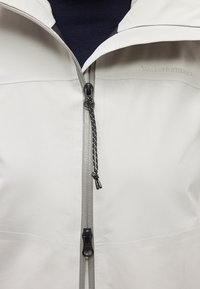 Κοντινό πλάνο φερμουάρ ενός ανοιχτόχρωμου γκρι τζάκετ με μαύρο κορδόνι έλξης, μερικώς ανοιχτό πάνω από βαθύ εσωτερικό στρώμα.