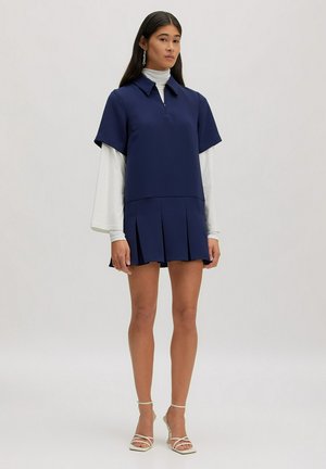 Jeune femme portant une robe bleu marine à manches courtes avec une jupe plissée par-dessus un col roulé blanc à manches longues, debout dans un espace neutre.