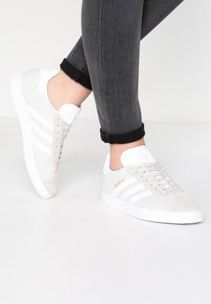 Baskets blanches Adidas Gazelle avec détails en mesh et suède portées avec un jean sombre retroussé sur un fond clair.
