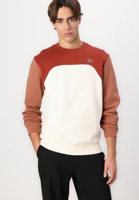 Genser med rund hals, hvit kropp og brune skuldre. Laget av myk stoff, har ribbestrikkede mansjetter og en liten logo på brystet.