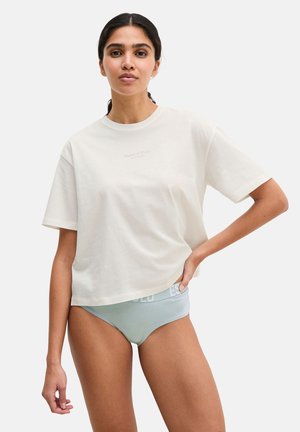 Vrouw draagt een oversized wit T-shirt en lichtblauwe ondergoed, staat met één hand op haar heup tegen een effen achtergrond.