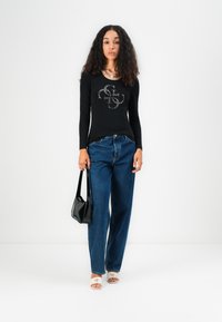 Svart langermet skjorte med grafisk design, blå høyhåndt denimjeans, svart håndveske og åpne hvite sandaler med branding.