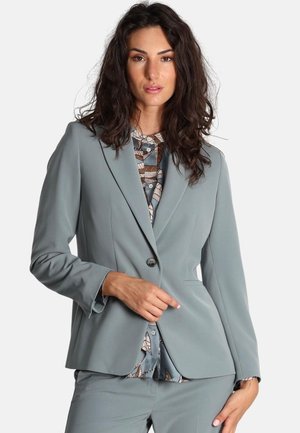 Blazer su misura grigio chiaro con revers, chiusura a un bottone e tasche laterali. Il tessuto ha una texture liscia; abbinato a una blouse a fantasia.