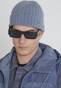 Siva rebrasta pletena kapa, crne kvadratne sunčane naočale i plava ženske jakne od kordura s kapuljačom, od teksturiranog materijala.