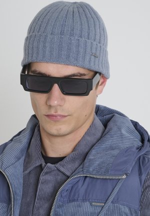 Siva rebrasta pletena kapa, crne kvadratne sunčane naočale i plava ženske jakne od kordura s kapuljačom, od teksturiranog materijala.