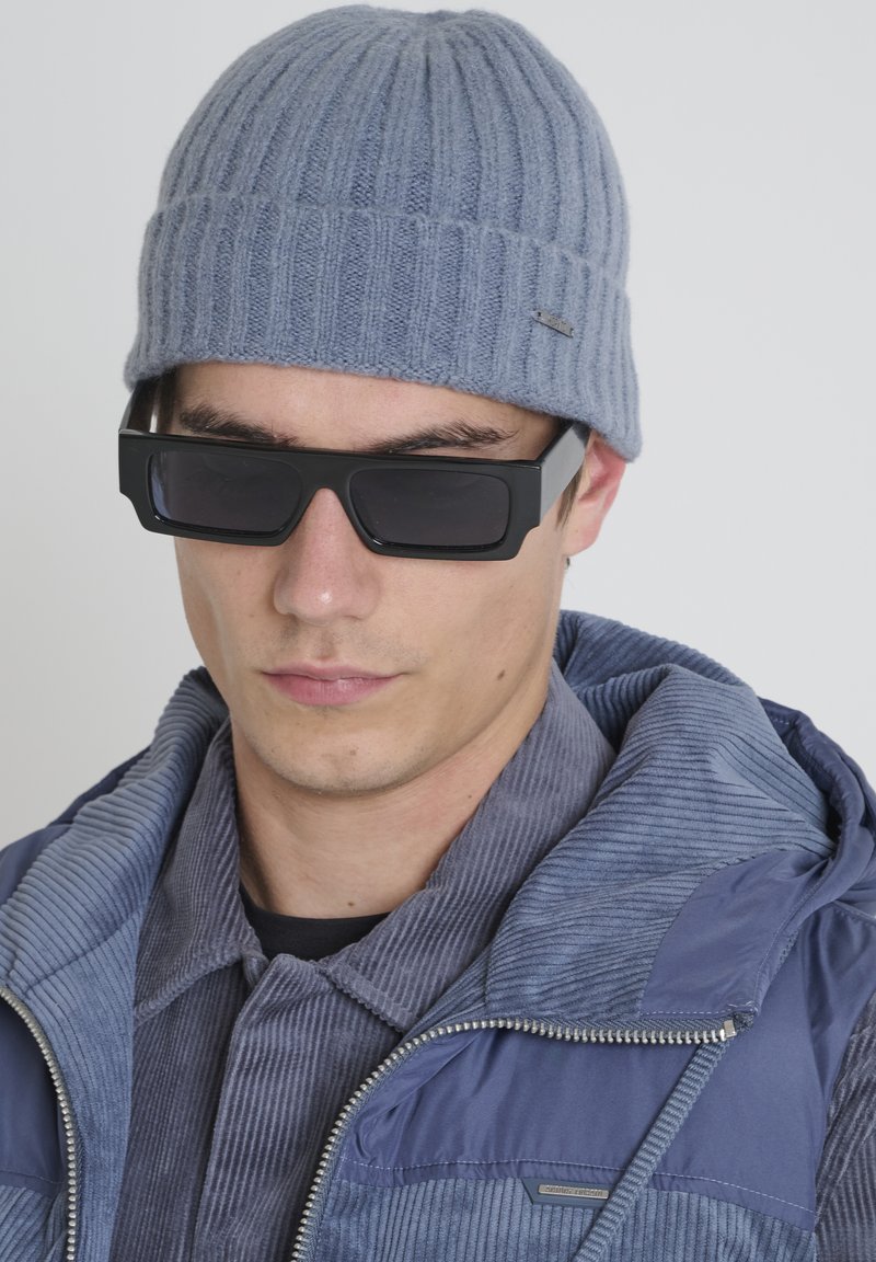 Siva rebrasta pletena kapa, crne kvadratne sunčane naočale i plava ženske jakne od kordura s kapuljačom, od teksturiranog materijala.