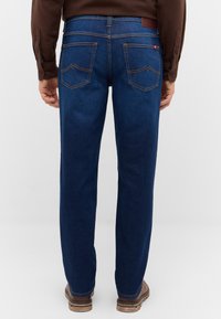 Ciemnoniebieskie dżinsy o prostej nogawce; mają dwie tylne kieszenie z detalami przeszyć oraz skórzaną metkę na pasku.