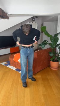 Persoon trekt een zwarte trui over het hoofd, staande op een houten vloer dichtbij een oranje bank en een grote potplant in de woonkamer.