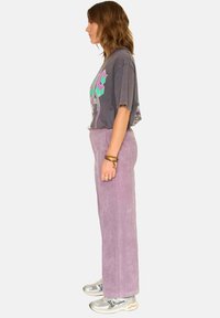 Pantalons larges en velours côtelé lavande au toucher doux, associés à un t-shirt graphique gris et des baskets argentées, vus de côté.