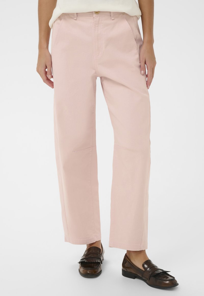 Személy világos rózsaszín egyenes szárú nadrágban és sötétbarna bőr mokaszinban, egyszínű háttér előtt állva.