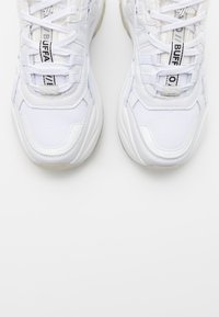 Bílé sportovní tenisky z kombinace síťoviny a syntetických materiálů, s kulatou špičkou, silnou podrážkou a kontrastními černými tkaničkami s logem.