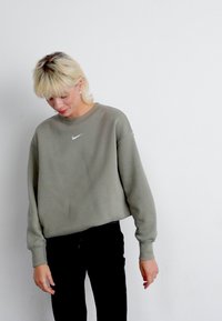 Sweatshirt Nike em verde oliva, feito de tecido macio com gola redonda. Apresenta um logo branco da Nike no peito. Combinado com calças pretas.