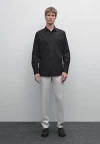 Schwarz-gestreiftes Langarmshirt mit Knopfverschluss, kombiniert mit hellbeigen Hosen und schwarzen Schuhen, vor einem einfarbigen Hintergrund.