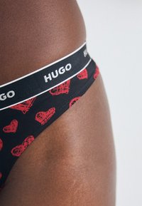 Czarne stringi z wzorem czerwonych serc, z czarnym pasem w talii z białym logo "HUGO". Gładka tekstura tkaniny.
