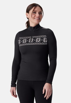 Femme souriante portant un haut noir à manches longues avec une fermeture éclair et un motif de flocons de neige blancs sur la poitrine et les épaules.