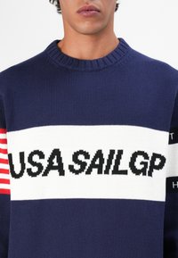 Navy knit sweater with a ribbed neckline; features a white horizontal stripe with "USA SAILGP" in bold black and red accent stripes on sleeves.