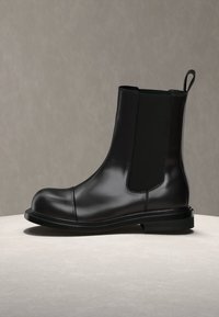 Λείο μαύρο δερμάτινο μποτάκι μέχρι τον αστράγαλο με ελαστικές πλευρικές πάνελ, στρογγυλεμένη μύτη, χαμηλό τακούνι και θηλιά στο πίσω μέρος, τοποθετημένο σε ουδέτερη επιφάνεια.