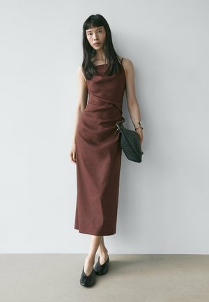 Donna in piedi contro il muro, indossa un vestito midi marrone senza maniche, scarpe nere, un braccialetto e tiene in mano una pochette intrecciata nera.