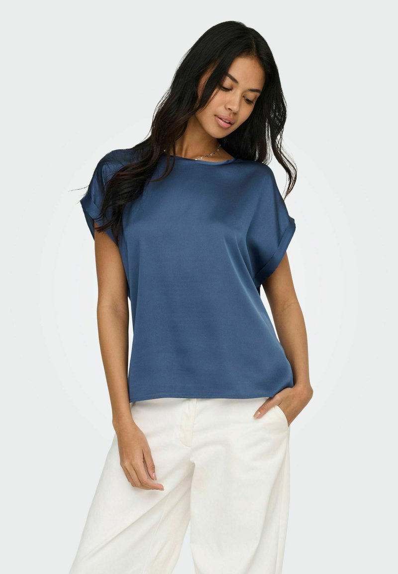 Femme aux longs cheveux foncés portant un chemisier bleu ample à manches courtes et un pantalon blanc, posant avec une main dans la poche sur fond blanc.