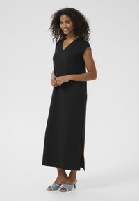 Robe maxi noire avec un décolleté en V, des manches courtes, des fentes sur les côtés et une texture lisse. Associée à des sandales à talons bleues.