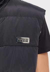 Gilet matelassé noir avec une étiquette de logo argentée brillante "Versace Jeans Couture", présentant une texture lisse et un design sans manches.