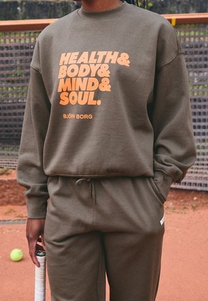 Person wearing brown tracksuit with "Health & Body & Mind & Soul" text on sweatshirt, holding a tennis racket on clay court.
