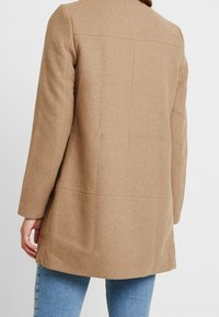 Hnědý vlněný kabát s volným střihem, který má detail zadního švu a dlouhé rukávy. Materiál má měkký, texturovaný povrch.