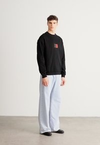 Μαύρο φούτερ με τετράγωνο γραφικό στο στήθος, σε συνδυασμό με ανοιχτό μπλε τζιν παντελόνι με φαρδιά μπατζάκια και μαύρα παπούτσια, στέκεται μπροστά σε ουδέτερο φόντο.