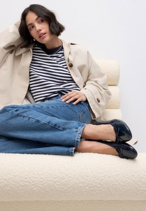 Mujer con abrigo beige, camiseta de rayas blancas y negras, vaqueros azules y zapatillas de ballet negras reclinada en un sofá de color crema con textura.