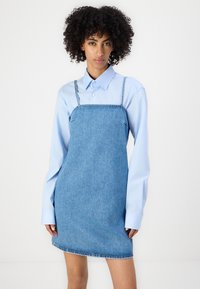 Kleid aus hellblauem Denim mit dünnen Trägern, das ein strukturiertes Design und eine Länge bis zur Mitte des Oberschenkels aufweist, kombiniert mit einem langärmeligen blauen Hemd.