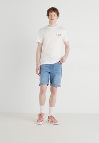 Λευκό κοντομάνικο μπλουζάκι με το λογότυπο "LEE", ανοιχτό μπλε τζιν σορτσάκι και ροζ αθλητικά παπούτσια. Το μοντέλο στέκεται σε ουδέτερο φόντο.