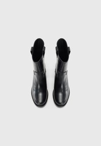 Une paire de bottes en cuir noir à la cheville avec des onglets de traction, finition lisse, bout rond et petits talons empilés sur fond blanc uni.