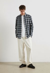 Karierter Flanellhemden in Marineblau, Grau und Weiß; weißes T-Shirt darunter; locker sitzende cremefarbene Hose; schwarze Schnürschuhe.