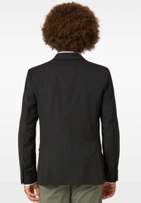 Melnas pielāgotas žaketes ar gludu tekstūru, ar vienu ventu aizmugurē un pogātām aprocēm. Kombinēta ar gaišām biksēm.