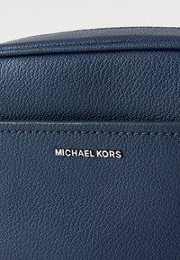 Torba iz teksturirane temno modre usnjene oblike z srebrnim logotipom »Michael Kors«. Ponaša se z zgornjim šivom in gladkim, zaobljenim dizajnom na zgornjem robu.