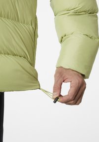 Chaqueta de plumas verde claro con una superficie texturizada, que presenta un cordón ajustable en el dobladillo, sostenida por una mano, mostrando utilidad y diseño.