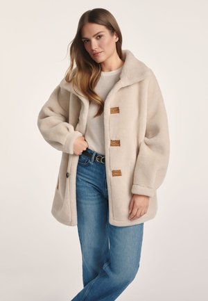 Kremowy polarowy płaszcz z szerokim kołnierzem, zapięciem na guziki typu toggle i bocznymi kieszeniami; zestawiony z jasnoniebieskimi dżinsami o prostym kroju.