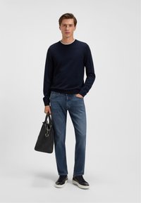 Navy crew-neck sweater, blue jeans, and black sneakers. Holding a textured black bag. Simple design, casual fit, and solid colors.