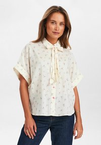Cream blouse with small pink floral pattern, short sleeves, and bow tie collar. Features pink buttons and rolled cuffs. Soft fabric texture.