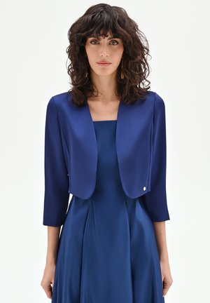 Donna con capelli ricci scuri che indossa un vestito blu senza maniche abbinato a una giacca corta blu con maniche a tre quarti, su sfondo bianco.