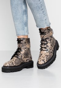 Bottines à motif peau de serpent en beige et noir, avec des lacets ronds, des œillets en métal et une semelle extérieure noire robuste.