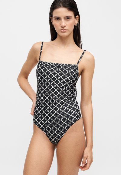 Femme portant un maillot de bain une pièce à motifs noir et blanc avec fines bretelles sur un fond clair uni.