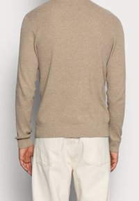 Suéter de punto beige con un patrón texturizado, mangas largas y puños acanalados, combinado con pantalones de color claro.