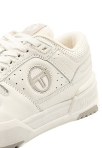 Bílé sportovní boty s perforovaným koženým svrškem, polstrovaným límcem, plochými tkaničkami a detaily loga na boku. Texturovaná gumová podešev.