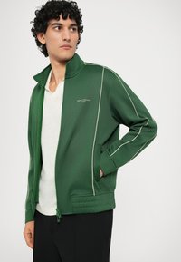 Green zip-up jacket with white piping, featuring a high collar, two side pockets, and a textured hem. Made of smooth fabric.