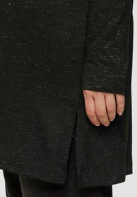 Langärmliges schwarzes Oberteil aus strukturiertem Stoff mit weißen Sprenkeln, seitlichem Schlitz und verlängerten Ärmeln. Eine Hand wurde auf den Stoff gelegt, um Details zu zeigen.