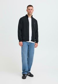Giacca nera con zip sopra una maglietta bianca, abbinata a jeans azzurri chiari e sneakers nere, in piedi davanti a uno sfondo bianco.