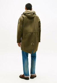 Wodoodporna kurtka w oliwkowo-zielonym kolorze z kapturem, zapięciem na zatrzaski i tylnym rozcięciem; w zestawie z niebieskimi dżinsami i brązowymi butami. Gładka faktura, długość do kolan.