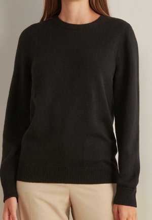 Frau trägt einen schlichten schwarzen Pullover mit langen Ärmeln und Rundhalsausschnitt, kombiniert mit beigen Hosen, und steht vor einem einfarbigen hellen Hintergrund.